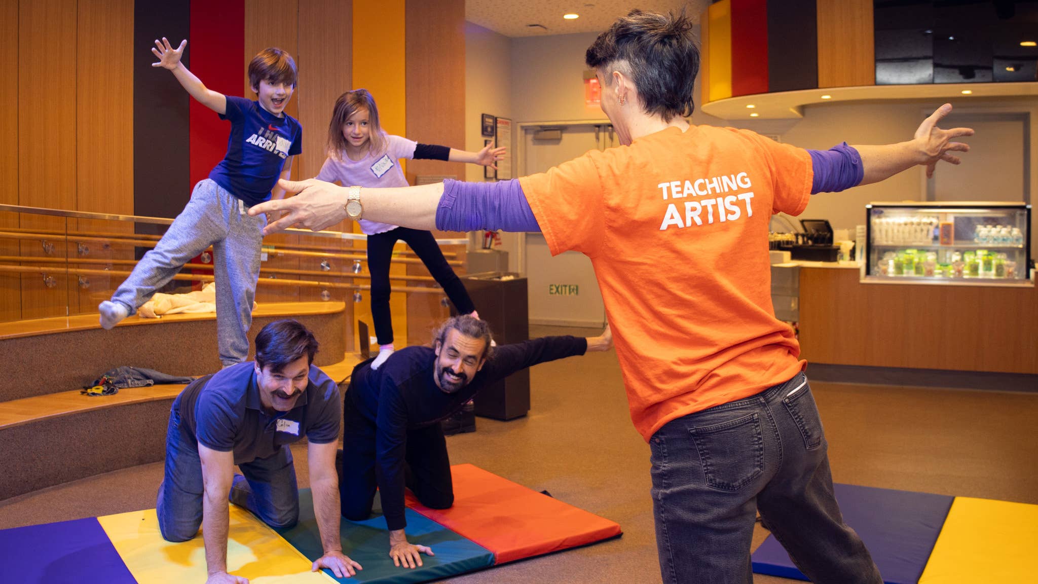 Instructor in orange “teaching artist” shirt leads children in a playful dance, arms out, in a community room.