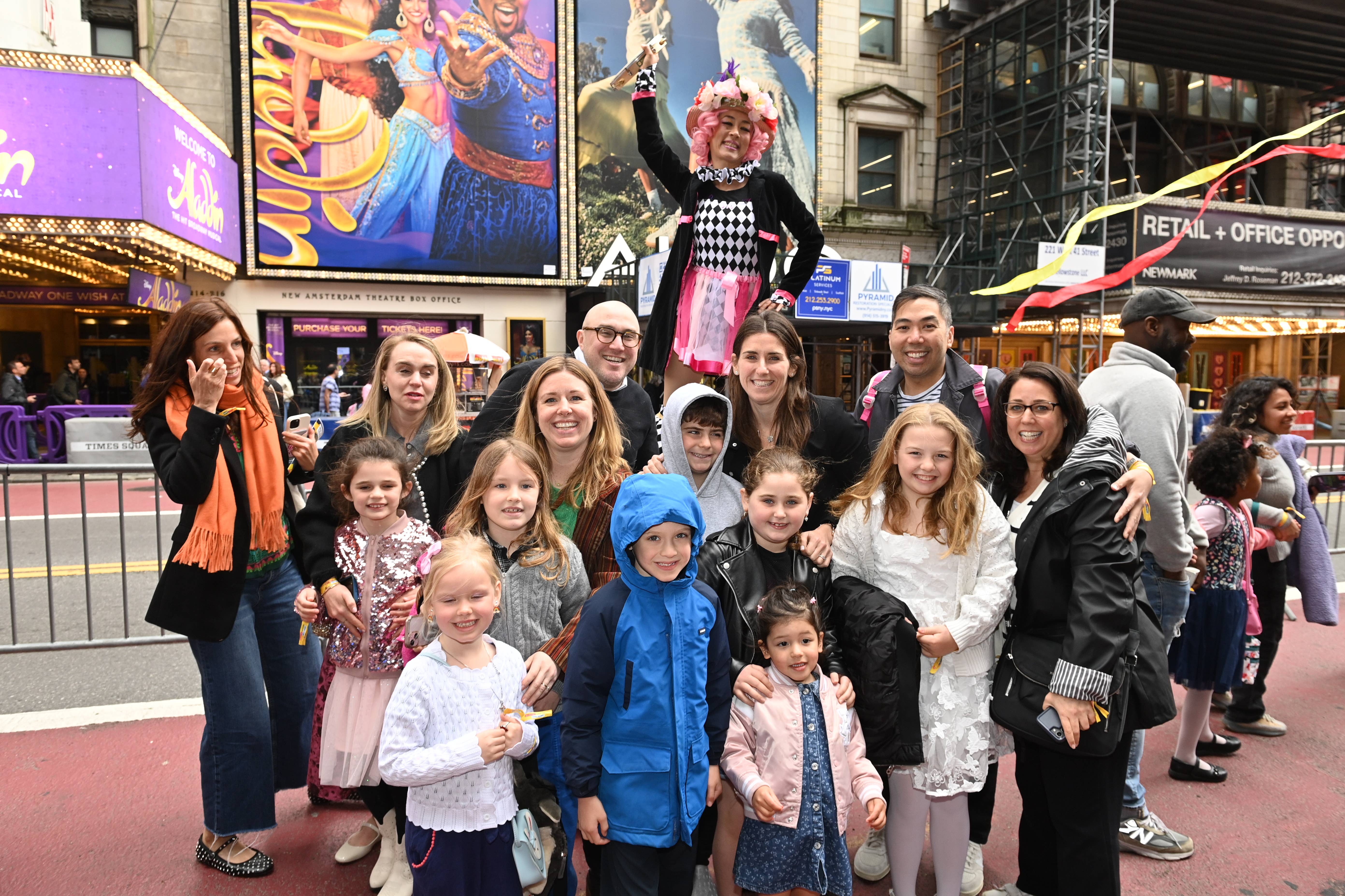 Costumed clown in pink and checkered outfit waves while families and children pose outside Broadway theater marquee