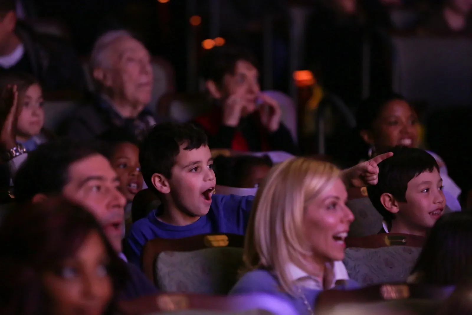 A group of kids and adults sitting in the audience look surprised and smile while watching a performance.