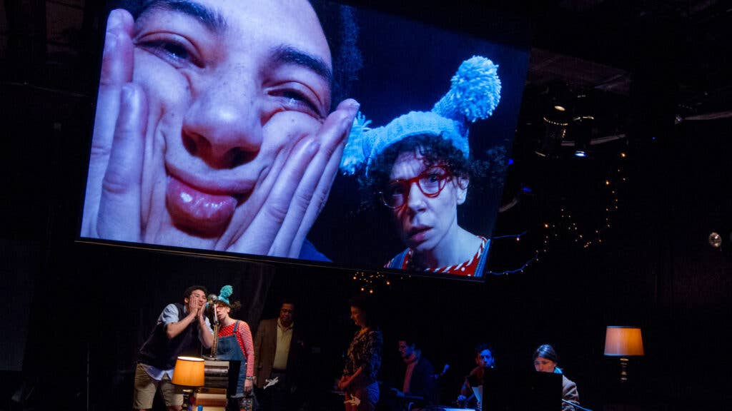 Two performers, one wearing a blue shirt and black vest and the other wearing a red shirt, overalls, red glasses and a blue hat, make silly faces on stage and appear on a large screen above the stage.