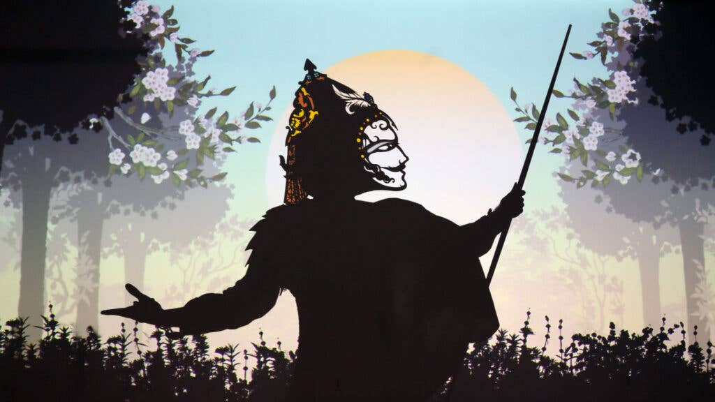 A performer wears a headpiece that casts a shadow of a face against a projection of trees and the sun.